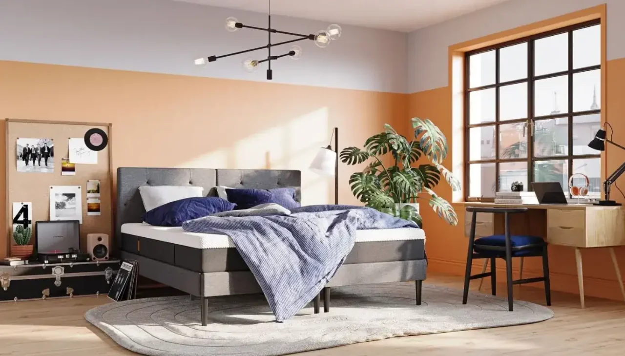 Um quarto com paredes em tons de laranja e branco, mobiliado com uma cama central que possui um colchão branco coberto por lençóis azuis.